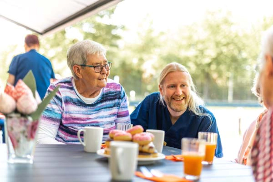 Patientin und Pflegekraft sitzen im Garten am Tisch mit mehreren Personen. Patientin schaut in die Fewrne zur Seite, Pflegekraft lacht eine weitere Person an.